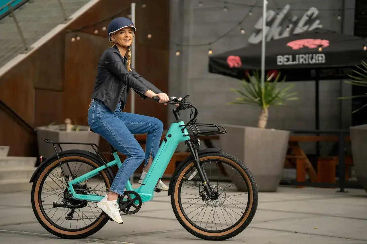 Uśmiechnięta kobieta w kasku i jeansach jedzie na turkusowym rowerze elektrycznym. To świetna okazja, by skorzystać z dofinansowania do roweru i cieszyć się jazdą!