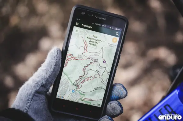 Rękawica trzyma smartfon z aplikacją **aplikacja trasy rowerowe** Trailforks, pokazującą mapę Beskidu Małego.